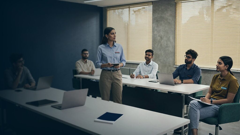 Woman standing with a tablet leading a meeting with new team members seated at white tables with laptops and notebooks in a modern office